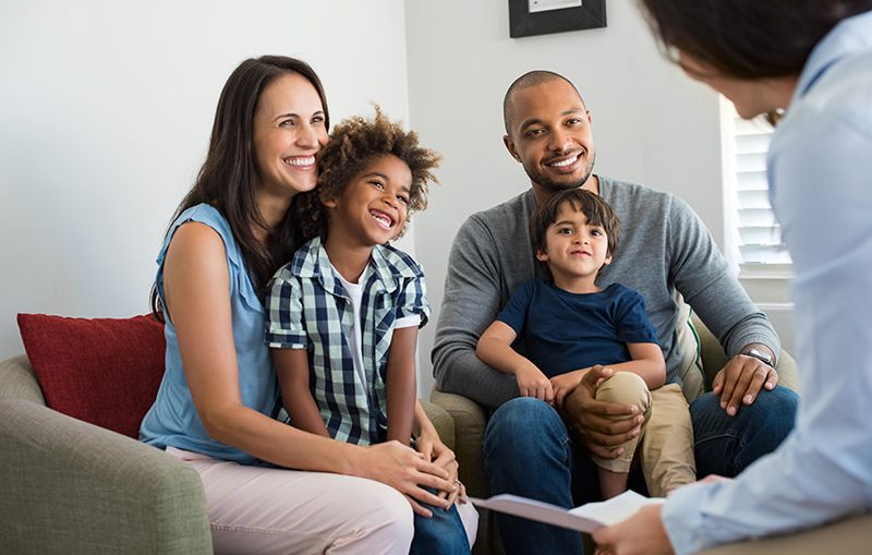 Happy young family sitting on couch and talking with family counselor. Smiling parents with adopted children discussing with counselor. Multiethnic family meeting a financial agent.